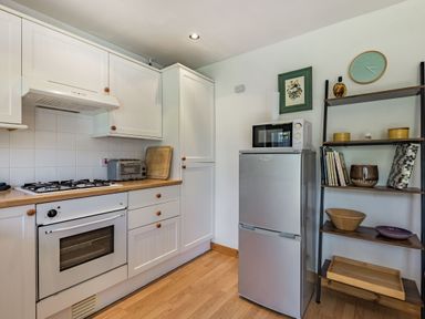 A kitchen with white cabinets a gas stove an oven a toaster a microwave on a silver refrigerator and a wooden shelving unit with books and decorative items at Gorwel in Llan Ffestiniog near Blaenau Ffestiniog