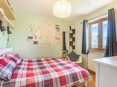 A bedroom with a bed covered in red plaid bedding a chair with a cushion a wall with maps and race bibs a window with curtains and a dresser at Gorwel in Llan Ffestiniog near Blaenau Ffestiniog