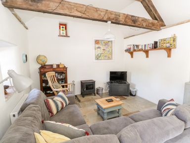 A living room with a large gray sectional sofa, wooden rocking chair, small coffee table, wood stove, TV on a stand, bookshelf, and exposed wooden beams at The Visiting House in Gorteen near Dunmore County Galway