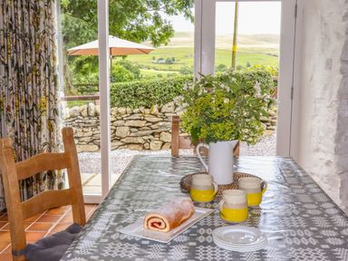 A dining table with a vase of flowers three cups plates and a cake near glass doors showing green fields at Y Bwthyn in Ysbyty Ifan