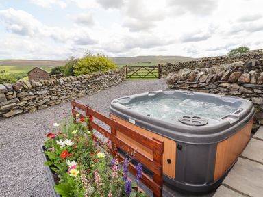 An outdoor hot tub next to a flower bed with a stone wall and green fields in the background at Y Bwthyn in Ysbyty Ifan