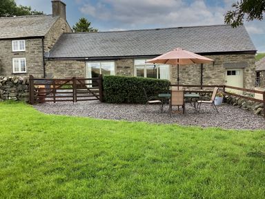 A stone house with a slate roof a small fenced patio with outdoor table chairs and umbrella and a green lawn at Y Bwthyn in Ysbyty Ifan