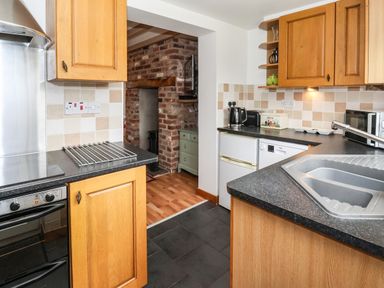 A kitchen with wooden cabinets a black countertop a stainless steel sink a stove kettle microwave and a tiled backsplash at The Cottage in Kilham near Driffield