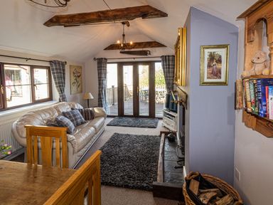 A living room with a beige sofa and gray cushions a wooden dining table a fireplace with a basket of firewood books on a wall shelf and glass doors leading to a deck at Cwm Derw Cottage in Llanafan Fawr near Builth Wells