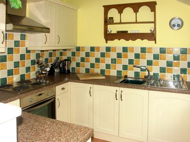 A kitchen with cream cabinets multicolored square tiled backsplash a stove oven sink and wooden shelf on yellow walls at Cwm Derw Cottage in Llanafan Fawr near Builth Wells