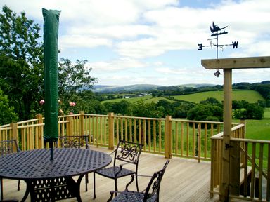 A wooden deck with metal table and chairs and a weather vane overlooking green fields and hills at Cwm Derw Cottage Llanafan Fawr near Builth Wells