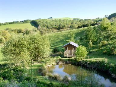 A small wooden cabin near a pond with trees and grassy hills at Cwm Derw Cottage Llanafan Fawr near Builth Wells
