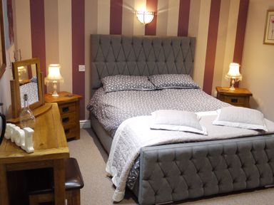 A bedroom with a double bed grey headboard striped wallpaper bedside tables with lamps and a wooden dressing table at Cwm Derw Cottage in Llanafan Fawr near Builth Wells