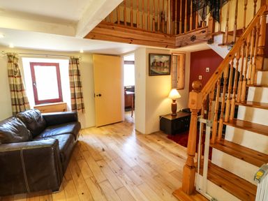 A living room with a brown leather sofa near a window with striped curtains and a wooden staircase with a lamp on a side table at Sutton Cottage in Ballysheen near Rosslare Harbour County Wexford