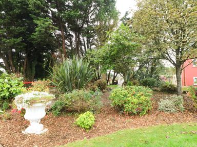 A garden with various bushes trees a white planter with flowers and a statue near a red building at Sutton Cottage in Ballysheen near Rosslare Harbour County Wexford
