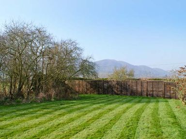 A garden with striped grass a wooden fence and bare trees with hills in the background at Brambles Cottage in Little Malvern near Malvern