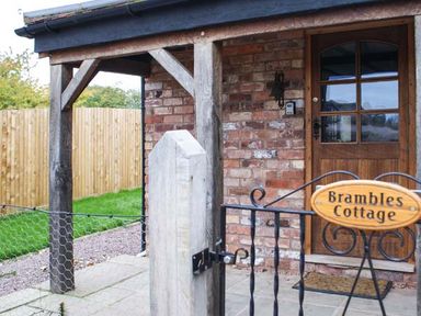 The entrance of a brick cottage with a wooden door and a metal gate with a sign reading Brambles Cottage at Little Malvern near Malvern
