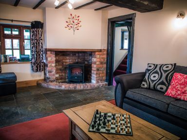 A living room with a brick fireplace and a wood stove a dark sofa with patterned cushions a wooden coffee table with a chessboard and a window with curtains at Brambles Cottage in Little Malvern near Malvern