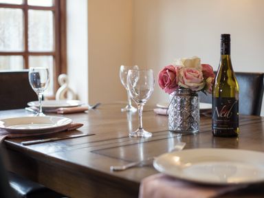 A dining table set with white plates pink napkins wine glasses a bottle of wine and a vase with roses at Brambles Cottage in Little Malvern near Malvern