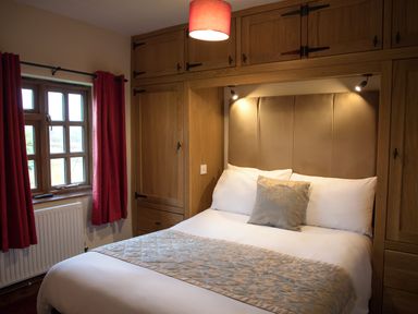 A bedroom with a double bed wooden overhead cabinets and a window with red curtains at Brambles Cottage in Little Malvern near Malvern