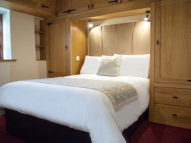 A bedroom with a large bed with white linens and a decorative pillow and bed runner surrounded by wooden built-in wardrobes at Brambles Cottage in Little Malvern near Malvern