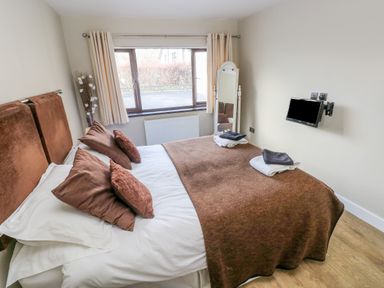 A bedroom with a double bed with brown pillows and blanket a floor mirror a wall mounted TV and a window at The Garden Flat in Saundersfoot