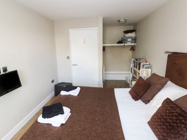 A bedroom with a bed made with brown and white bedding a wall-mounted television a small bookshelf and an open closet at The Garden Flat in Saundersfoot
