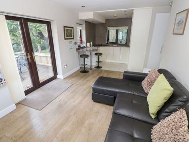 An open living room and kitchen area with a black leather sofa with cushions wooden floor a small bar with stools and double glass doors leading to an outdoor deck at The Garden Flat in Saundersfoot