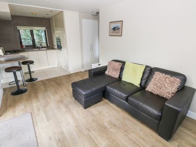 A living area with a black sectional sofa with three cushions and a small kitchen with bar stools at The Garden Flat in Saundersfoot