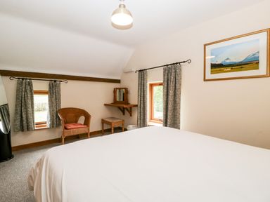 A bedroom with a bed white curtains a wicker chair a small dressing table and a framed landscape picture at Stable End in Luntley near Pembridge