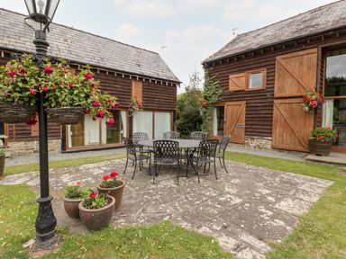 A patio area with a round table and chairs outside wooden buildings with flower pots and hanging baskets at Stable End in Luntley near Pembridge