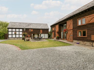 A courtyard with gravel and grass surrounded by timber and brick buildings with outdoor seating and hanging flower baskets at Stable End in Luntley near Pembridge