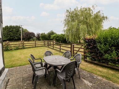 An outdoor patio area with a round table and six chairs next to a grassy yard with a tree and wooden fence at Stable End in Luntley near Pembridge