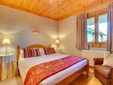 A bedroom with a wooden bed with striped pillows two bedside tables with lamps a window with curtains and a brown armchair at Apple Tree Lodge Swarland near Alnwick