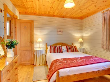 A bedroom with a wooden bed and red pillows and blanket two bedside tables with lamps a wooden dresser with a flower vase and a wooden door at Apple Tree Lodge Swarland near Alnwick