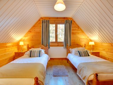 A bedroom with two single beds with striped pillows a window with curtains bedside tables with lamps and wooden walls at Apple Tree Lodge in Swarland near Alnwick