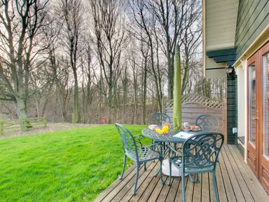 A patio with a metal table and four chairs set with breakfast items next to a grassy yard and leafless trees at Apple Tree Lodge Swarland near Alnwick