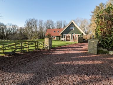 A house with a red roof and green exterior surrounded by grass trees and a gravel driveway at Lime Tree Lodge Swarland near Alnwick