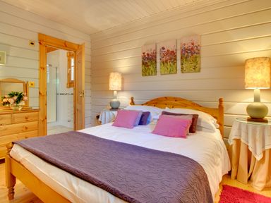 A bedroom with a wooden bed dressed with pillows and a blanket two bedside tables with lamps and a dresser with a mirror and flowers at Lime Tree Lodge in Swarland near Alnwick