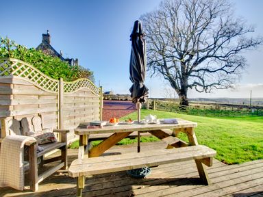 An outdoor wooden picnic table with an umbrella and bench on a deck near a wooden chair with a blanket and cushion and a tree in the background at Lime Tree Lodge in Swarland near Alnwick
