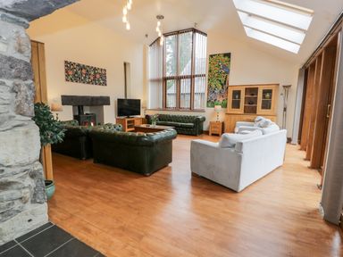 A living room with green and white sofas a wooden cabinet a stone fireplace and large windows at Ddol Helyg Barn in Llanrug