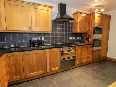 A kitchen with wooden cabinets black countertop black tiled backsplash built in oven and microwave at Ddol Helyg Barn in Llanrug