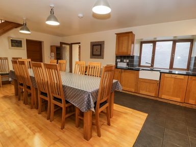 A kitchen and dining area with a large wooden table and chairs covered with a polka dot tablecloth and wooden cabinets under a window at Ddol Helyg Barn in Llanrug