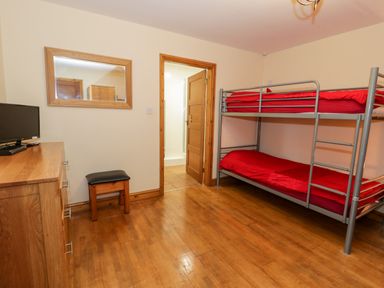 A bedroom with bunk beds with red bedding a wooden dresser a small stool a wall mirror and an open door to a bathroom at Ddol Helyg Barn in Llanrug