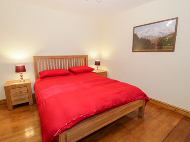 A bedroom with a wooden double bed with red bedding two wooden bedside tables each with a lamp and a framed landscape picture on the wall at Ddol Helyg Barn in Llanrug