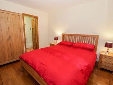 A bedroom with a wooden bed with red bedding two bedside tables with lamps a wooden wardrobe and an open door to a bathroom at Ddol Helyg Barn in Llanrug