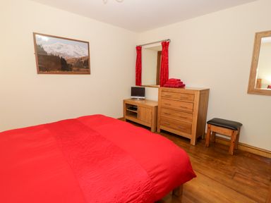 A bedroom with a red bedspread wooden dresser a small TV a wooden stool and a window with red curtains at Ddol Helyg Barn in Llanrug