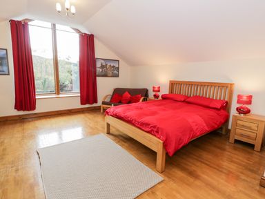 A bedroom with a wooden bed with red bedding a gray rug two wooden nightstands with red lamps a window with red curtains and a small sofa with red cushions at Ddol Helyg Barn in Llanrug