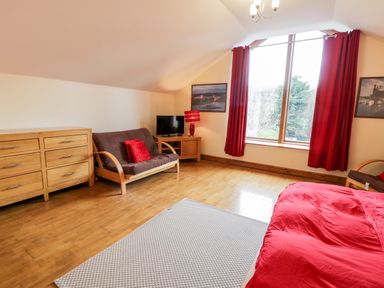 A room with a wooden dresser a futon with red cushions a small TV on a wooden stand red curtains on a window and a bed with red bedding at Ddol Helyg Barn in Llanrug