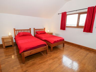 A bedroom with two single wooden beds with red bedding two wooden bedside tables with lamps and a window with red curtains at Ddol Helyg Barn in Llanrug