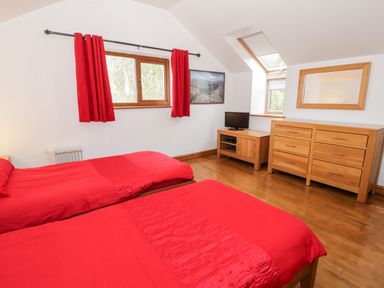 A bedroom with two red beds a wooden dresser a TV on a wooden stand and red curtains at Ddol Helyg Barn in Llanrug