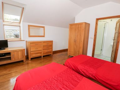 A bedroom with two single beds with red bedding a wooden wardrobe a chest of drawers a TV stand with a TV and a bathroom with a shower visible at Ddol Helyg Barn in Llanrug