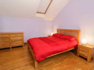 A bedroom with a wooden bed with red bedding two bedside tables with lamps and a chest of drawers at Ddol Helyg Barn in Llanrug