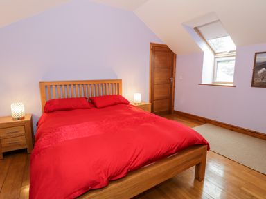 A bedroom with a wooden bed frame and red bedding two bedside tables with lamps a wooden door and a window with a blind at Ddol Helyg Barn in Llanrug