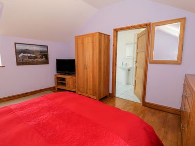 A bedroom with red bedspread wooden furniture television and an open door to a bathroom at Ddol Helyg Barn in Llanrug